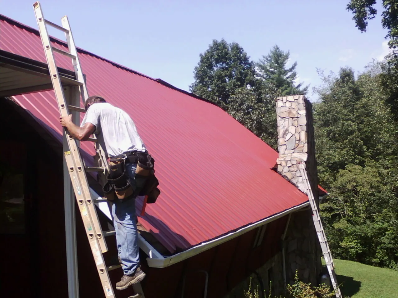 Roofer on a ladder beside a red metal roof and stone chimney — Soffit & Fascia Repair by Roofing Brothers in Horseshoe Bay